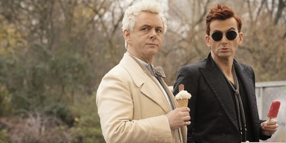 Two men, one dressed in all white and the other in all black, stand in a park eating ice cream.
