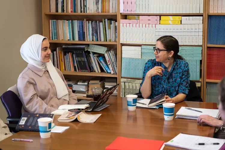 A student and professor sit at a table together in front of a bookshelf