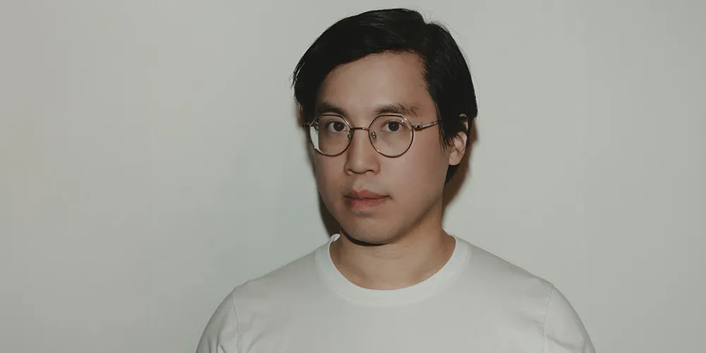 writer Tony Tulathimutte, man with brown hair in white t-shirt standing in front of white wall