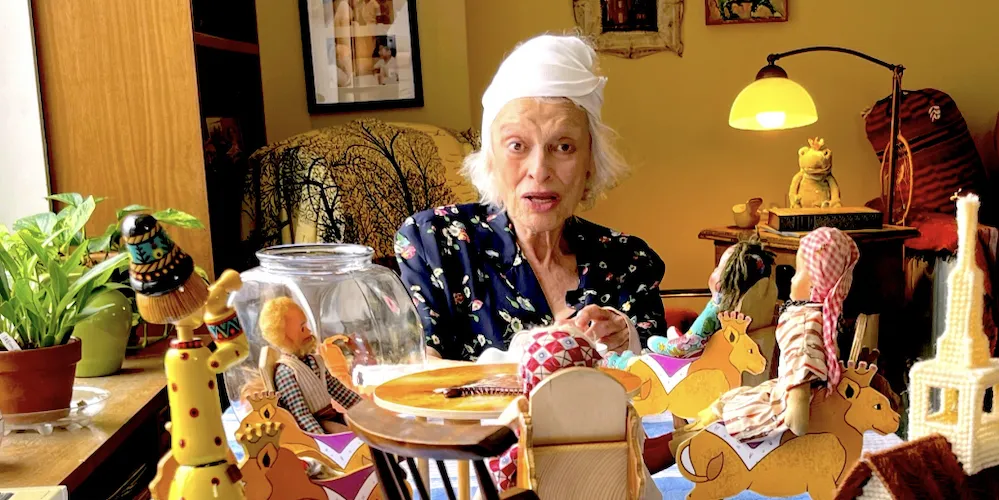An elderly woman sits at a table covered in toys and dolls