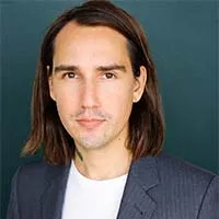 man with shoulder length brown hair wearing a white tshirt and blue blazer