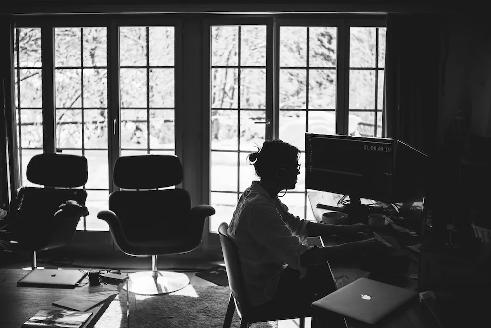 A silhouette of a woman sitting at a desk with computer monitors, in front of a window. 