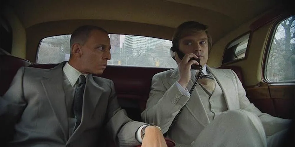 Two men in suits sit in a car, one on a car phone. 