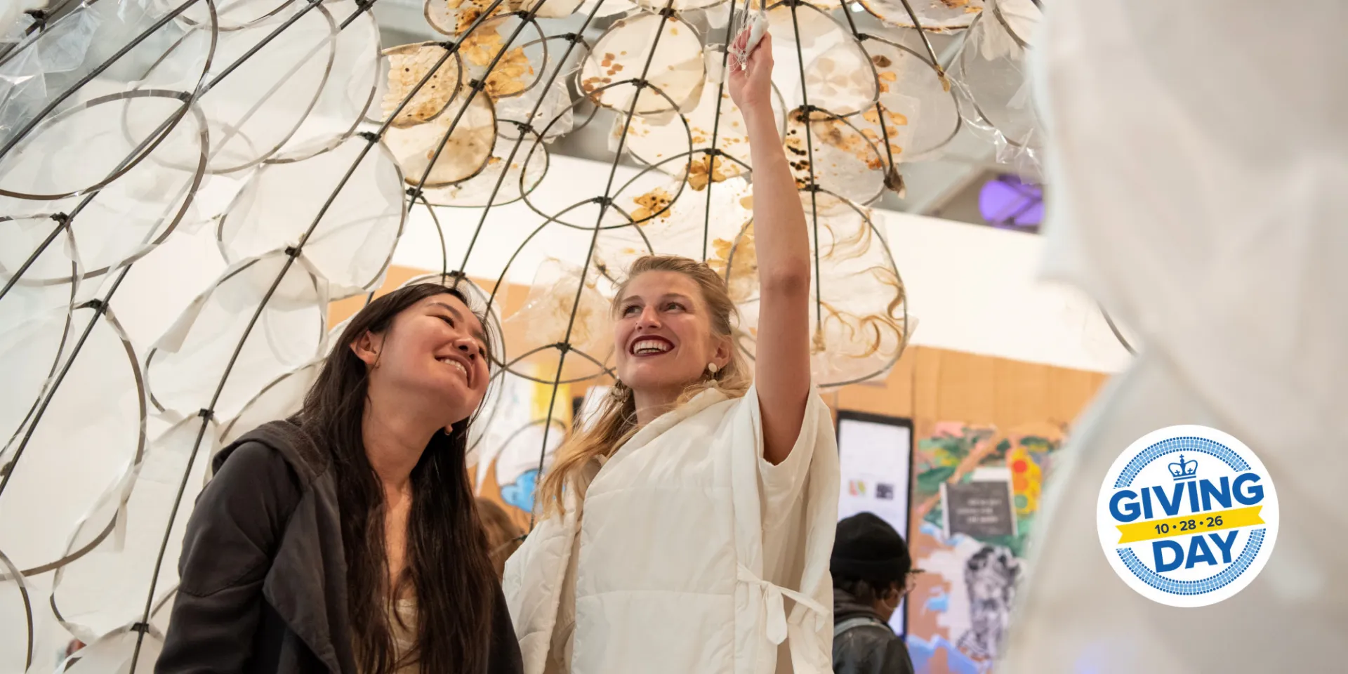 Two women stand under a visual arts sculpture, smiling.
