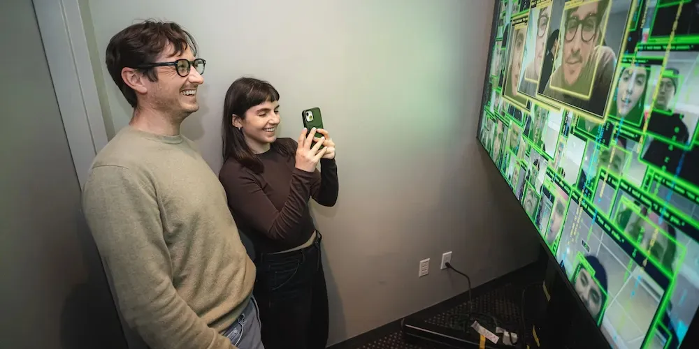 Two people stand in front of a screen with different images of their faces on it. 
