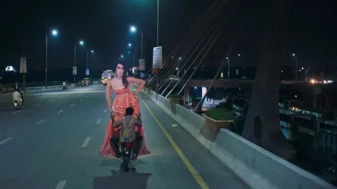 A man carries an oversized Cardboard figure of a trans woman performer on a motorbike