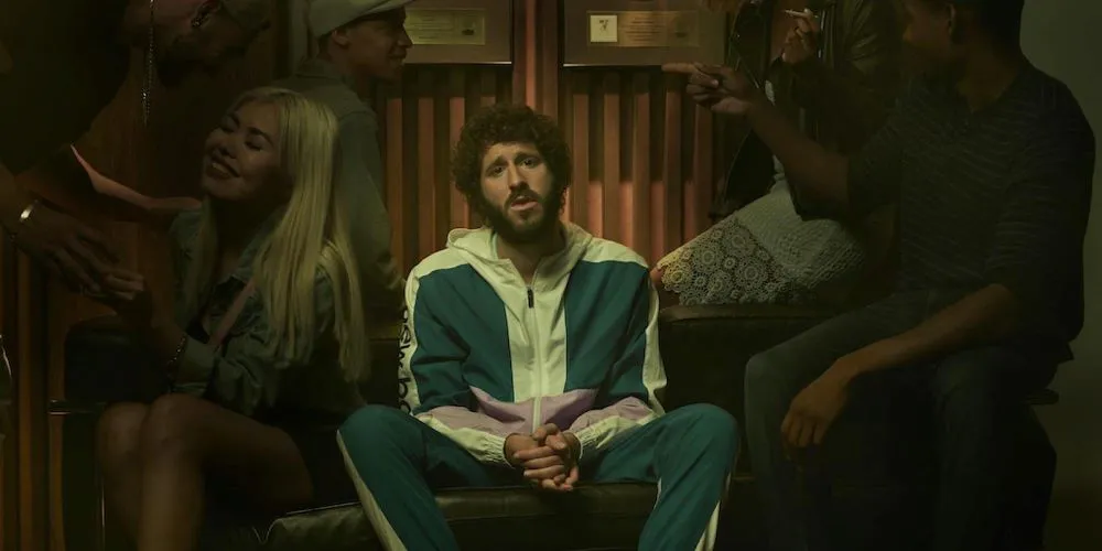 Still from Dave. Dave sits in the middle of a party.
