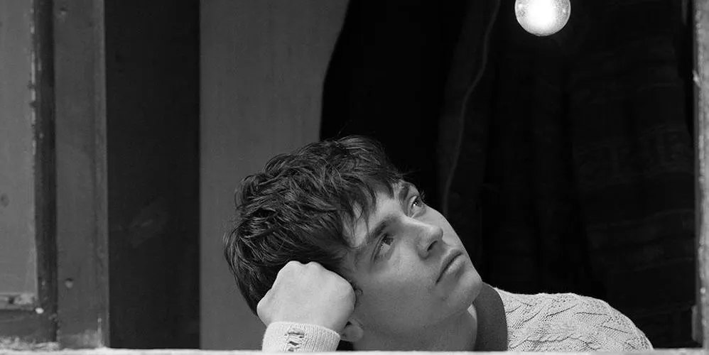 A young man leans on a windowsill and looks pensively at a lightbulb.