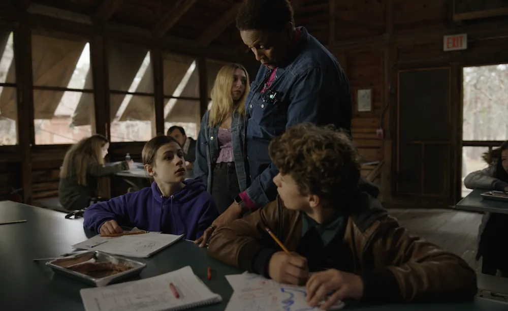 Still from Sound to Sea - Two children sit at a table in a cabin. A woman looks over their shoulders at their books laid out in front of them.
