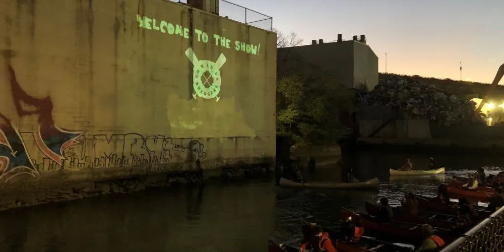 Graffitied concrete wall with a river full of people in canoes beneath it