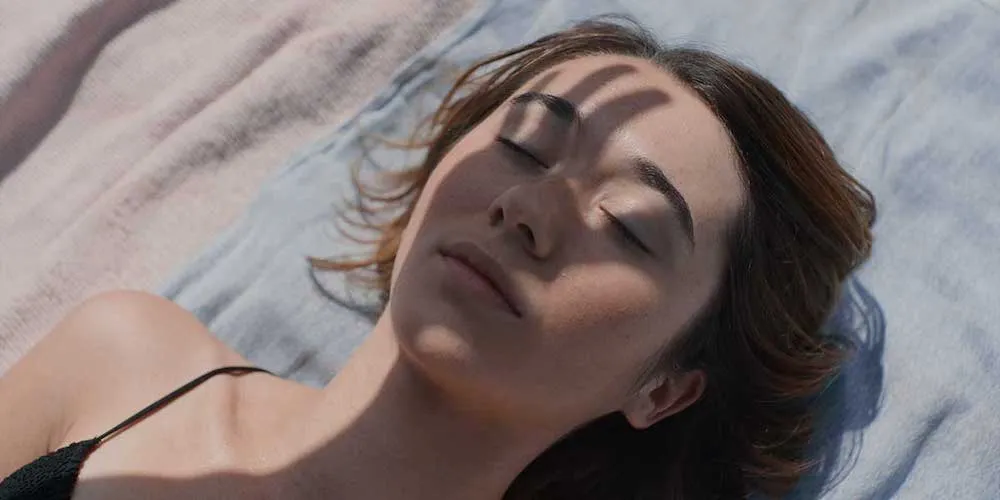 A woman lies in the sun with her eyes closed, the shadow of a hand falling across her face
