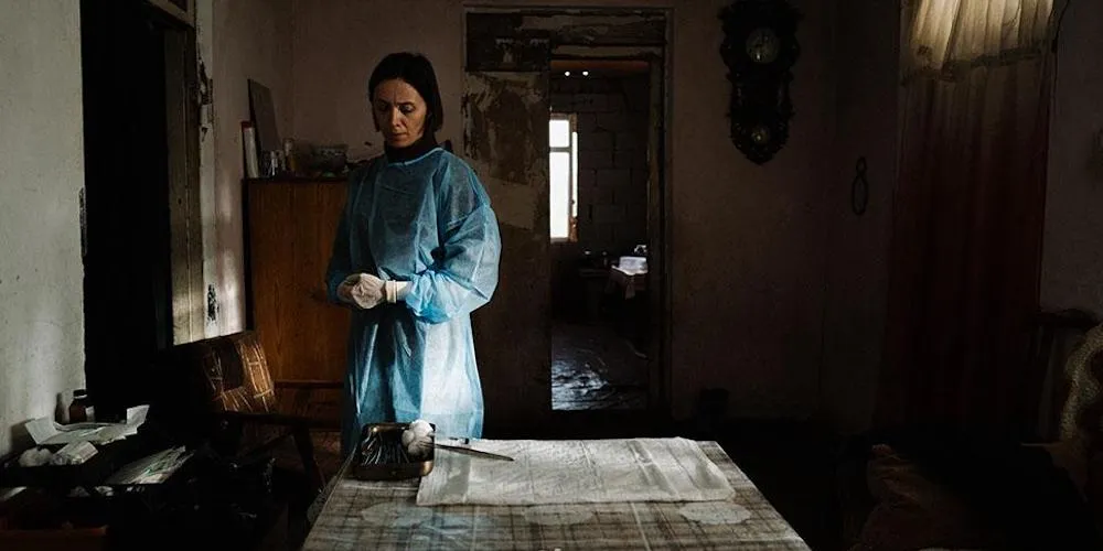 A person in a medical smock stands behind a kitchen table with medical tools. 