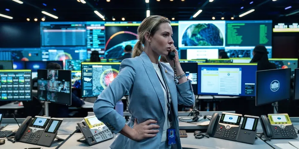 a woman stands in front of many computer screens, talking on the phone