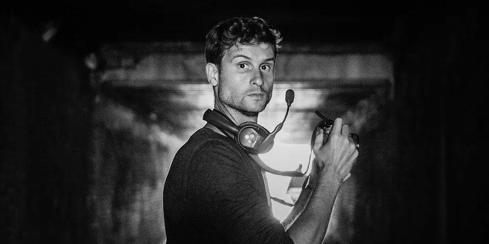 Black and white photo of man with headset around his neck standing in dark space backlit by a bright light tunneling in. 