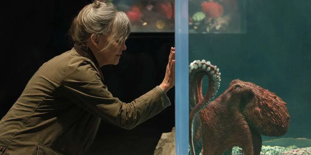 a woman holds her hand up to a glass aquarium with an octopus