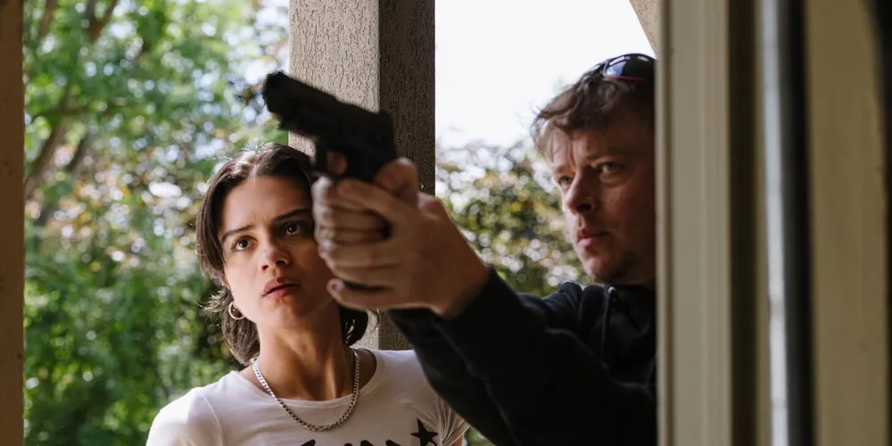 Two people stand behind a door, one holds a gun. 