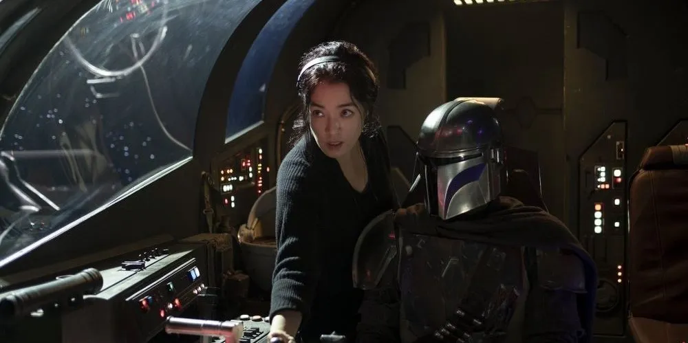Two characters in the cockpit of a spaceship, one is seated and wearing a helmet