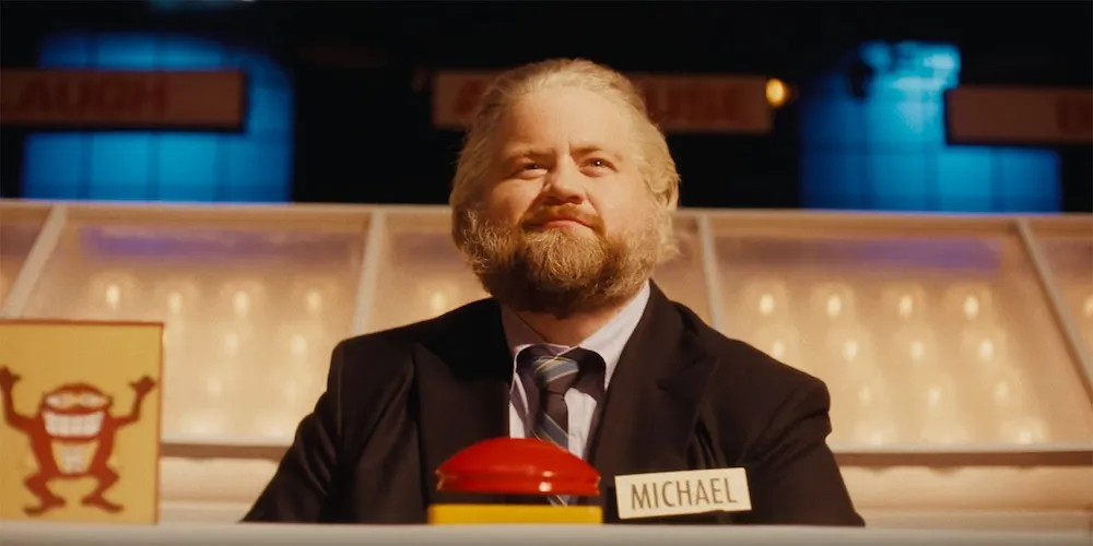 Man in suit sitting behind a large red gameshow buzzer. 