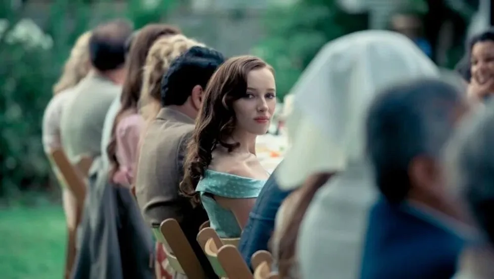 Woman sits at a long outdoor table and looks on at camera. 