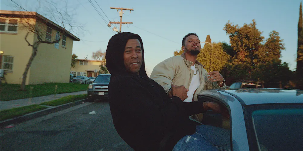 Two men hang out of a car window 