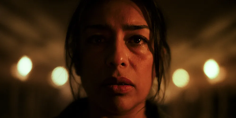 Close-up of a woman's face lit warmly, staring ahead with blurred lights behind. 
