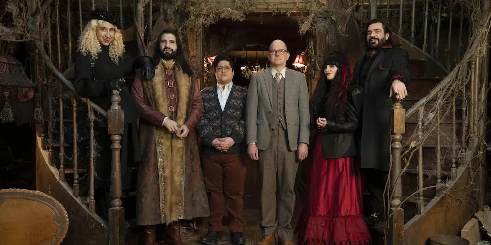 A group of people in fantastical clothing pose at the bottom of a grand staircase. 