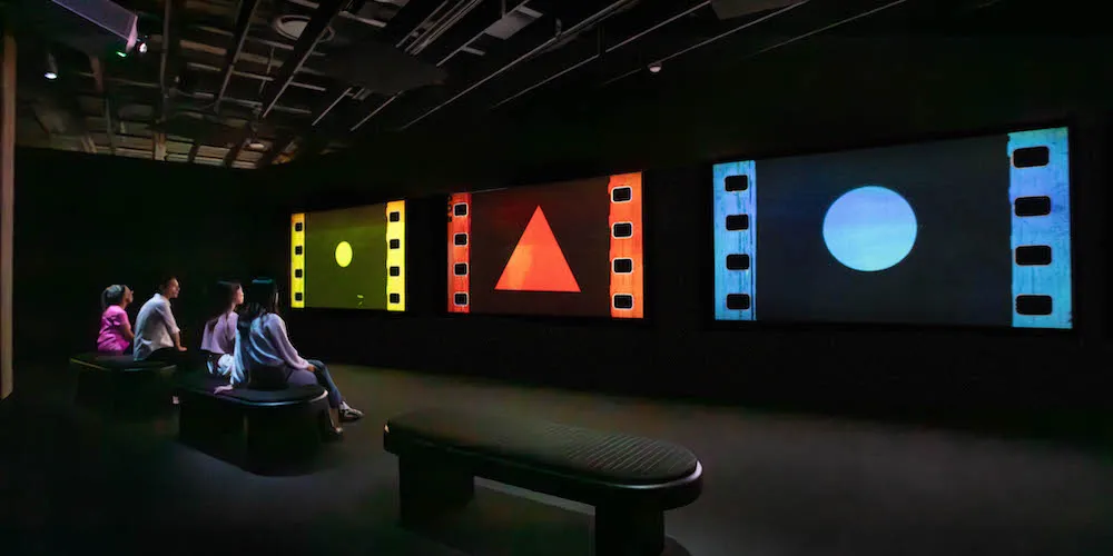 Viewers sitting on a bench in a dark gallery looking at three screens displaying a yellow circle, red triangle, and a blue dot. 