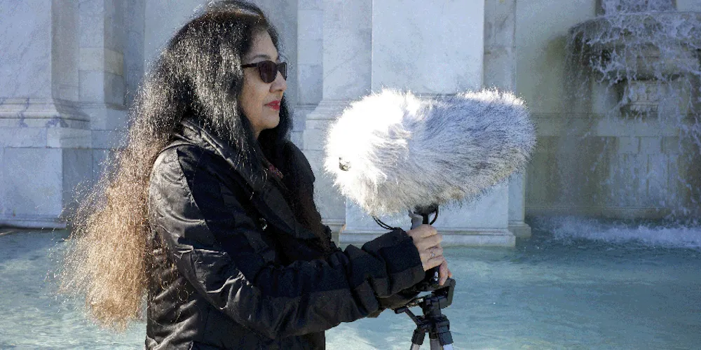 A woman holds a large, fluffy microphone in front of a fountain. 