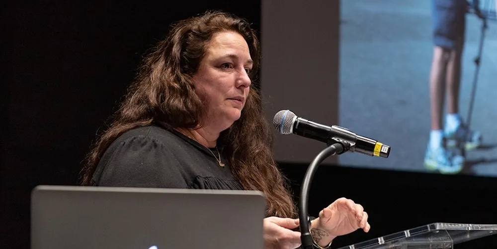 A woman speaks at a podium with an image projected behind her, just visible in the corner.