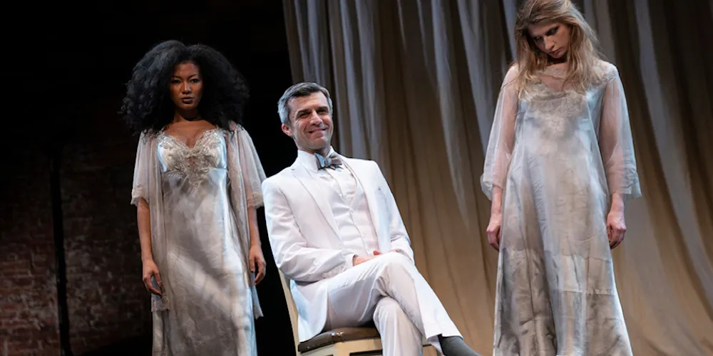 Two women standing and one man sitting in a chair on stage.