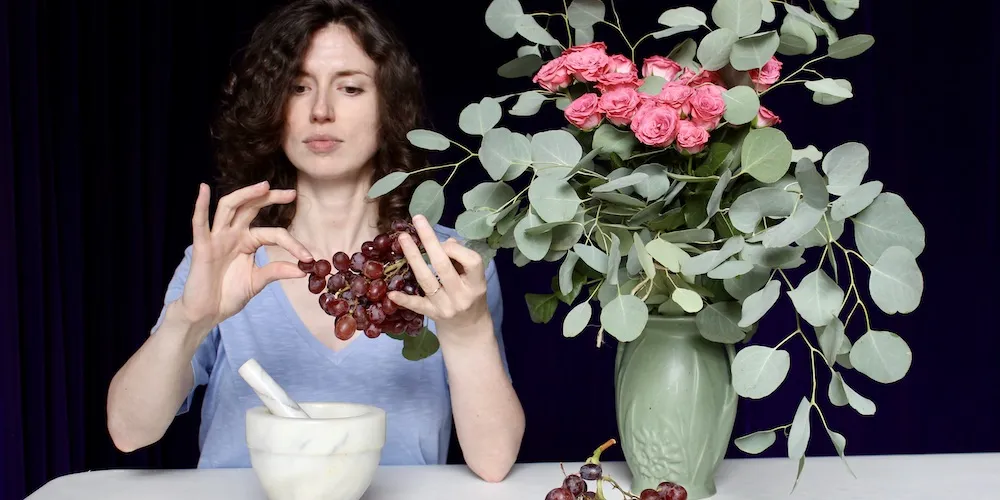 A woman sits at a table next to a vase of flowers and a mortar & pestle. She picks red grapes from a bunch. 
