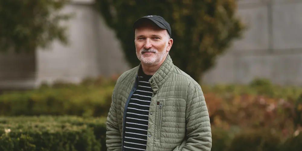 A man in a hat and green jacket stands outside, with greenery in the background. 