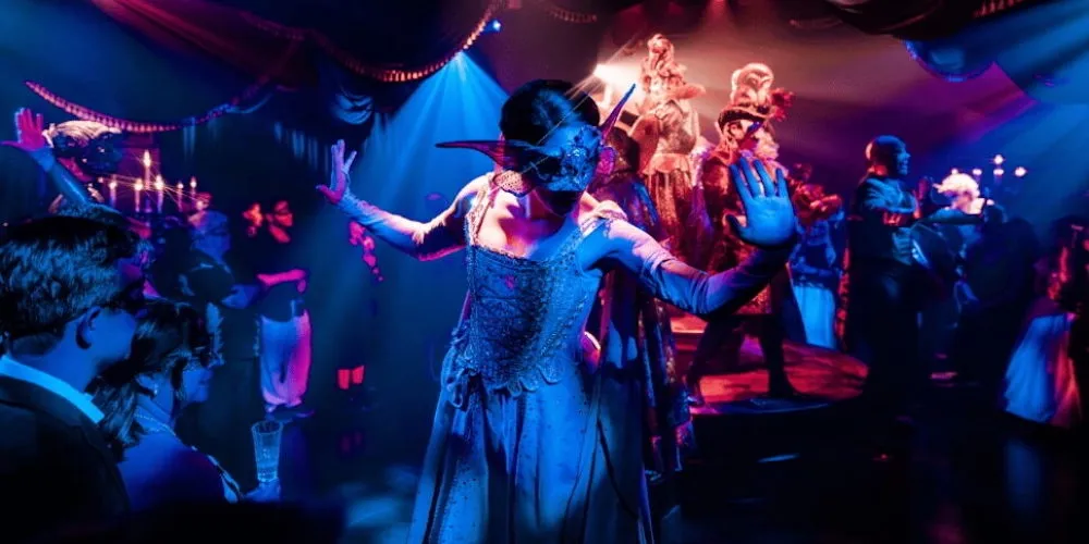 A woman in a mask stands with her arms outstretched. 