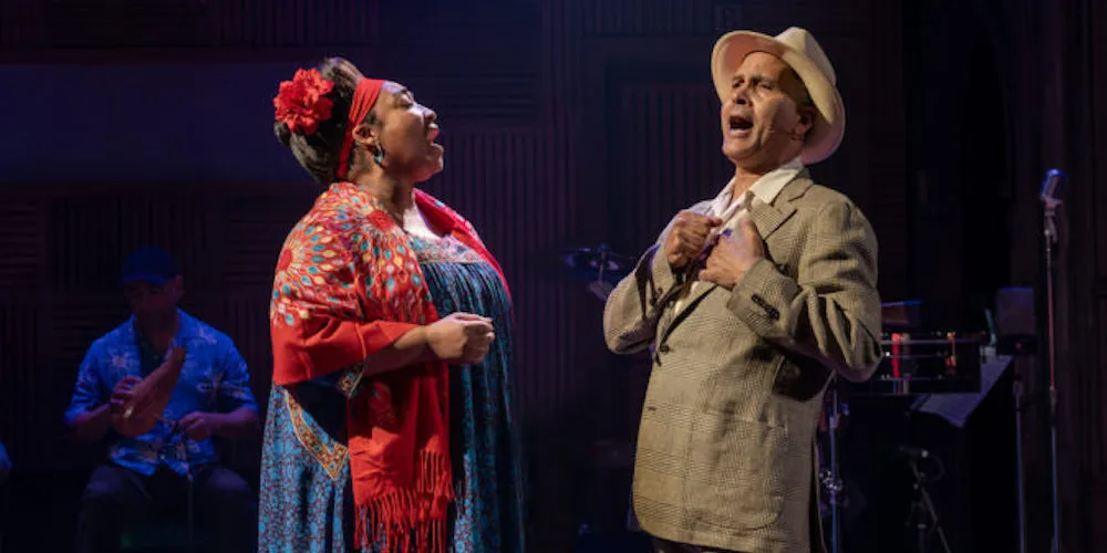 A woman in red and blue dress sings with a man in tan suit and hat on a stage.