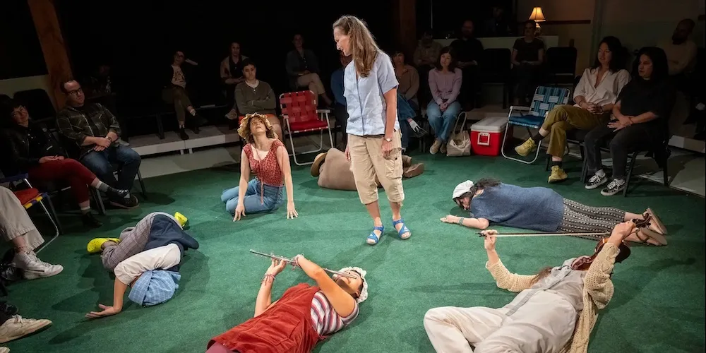 
A woman stands at the center of a circular stage, surrounded by actors who roll and move fluidly around her,