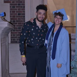 Two people stand outside a building, one in a cap and gown. 
