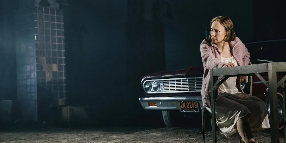 A woman sits at the table in front of a parked car.