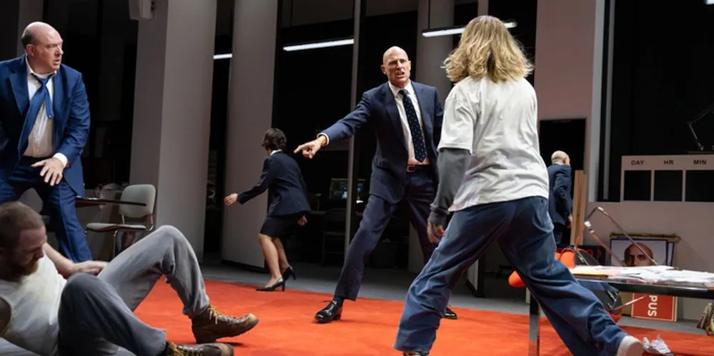 Actors perform a dynamic scene on stage with one performer seated on the floor as others move across the space.