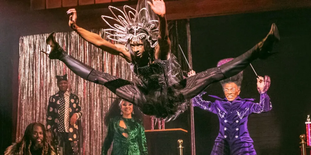 Dancer in a black costume split jumps front of group of actors. 