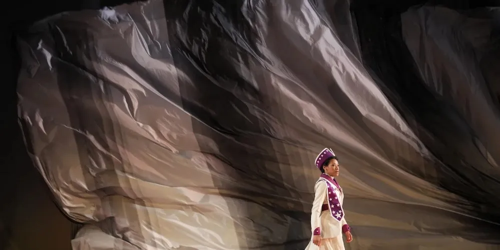 Woman walks across stage in front of giant grey flag flowing in the background.