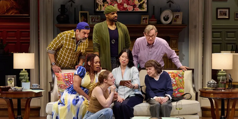 People sit on and stand around a couch looking at a phone. 