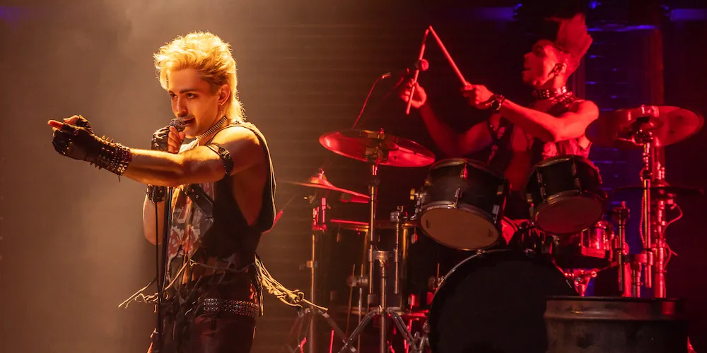 Man sings on stage with drummer in background.