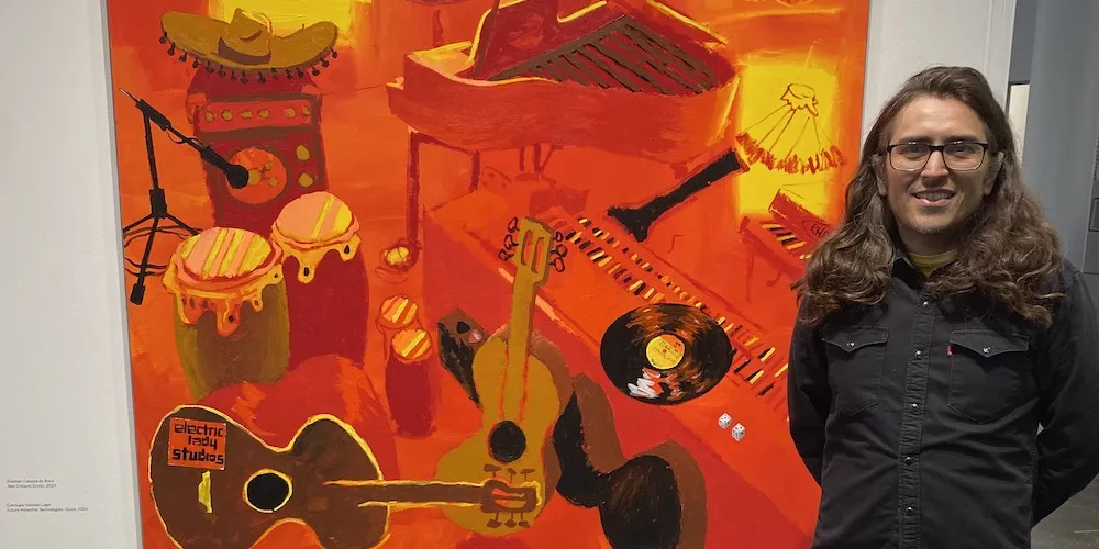 Man with long brown hairs stands next to orange and black painting of instruments. 