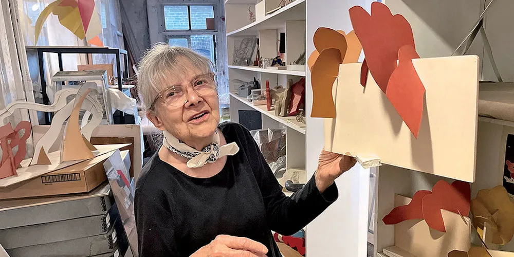 A woman stands in a studio, showing a piece of art to the camera. 
