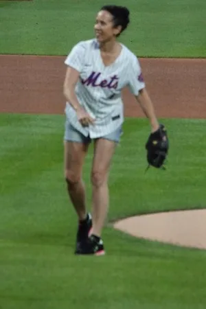 Woman in baseball jersey on the pitcher's mound of a baseball field throwing a baseball. 