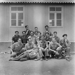Old black and white photograph of a group on men sitting in front of prison structure.