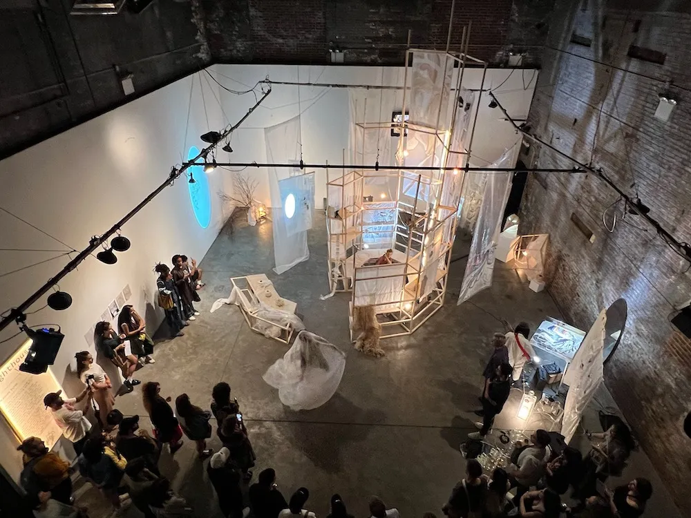 Ariel view of many people viewing an art installation featuring a structure made out of light wood and transparent materials with projections on the materials and surrounding walls. 