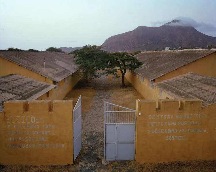 Yellow brick buildings with corrogated roofs behind prison wall with mountains in background.