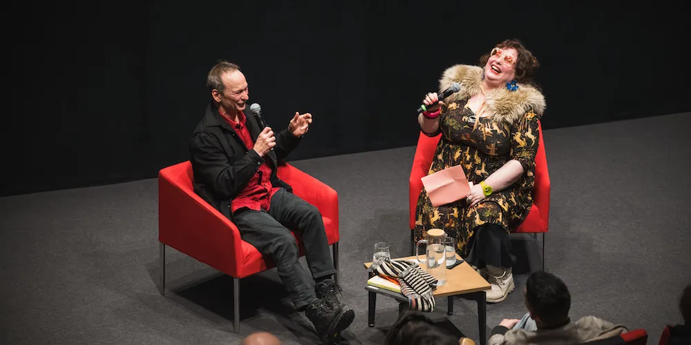 two people using microphones sit in red chairs, having a lively conversation