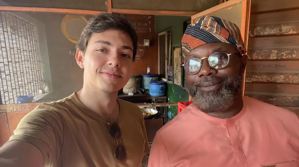Young man in brown t-shirt stands next to man with patterned hat in canteen. 